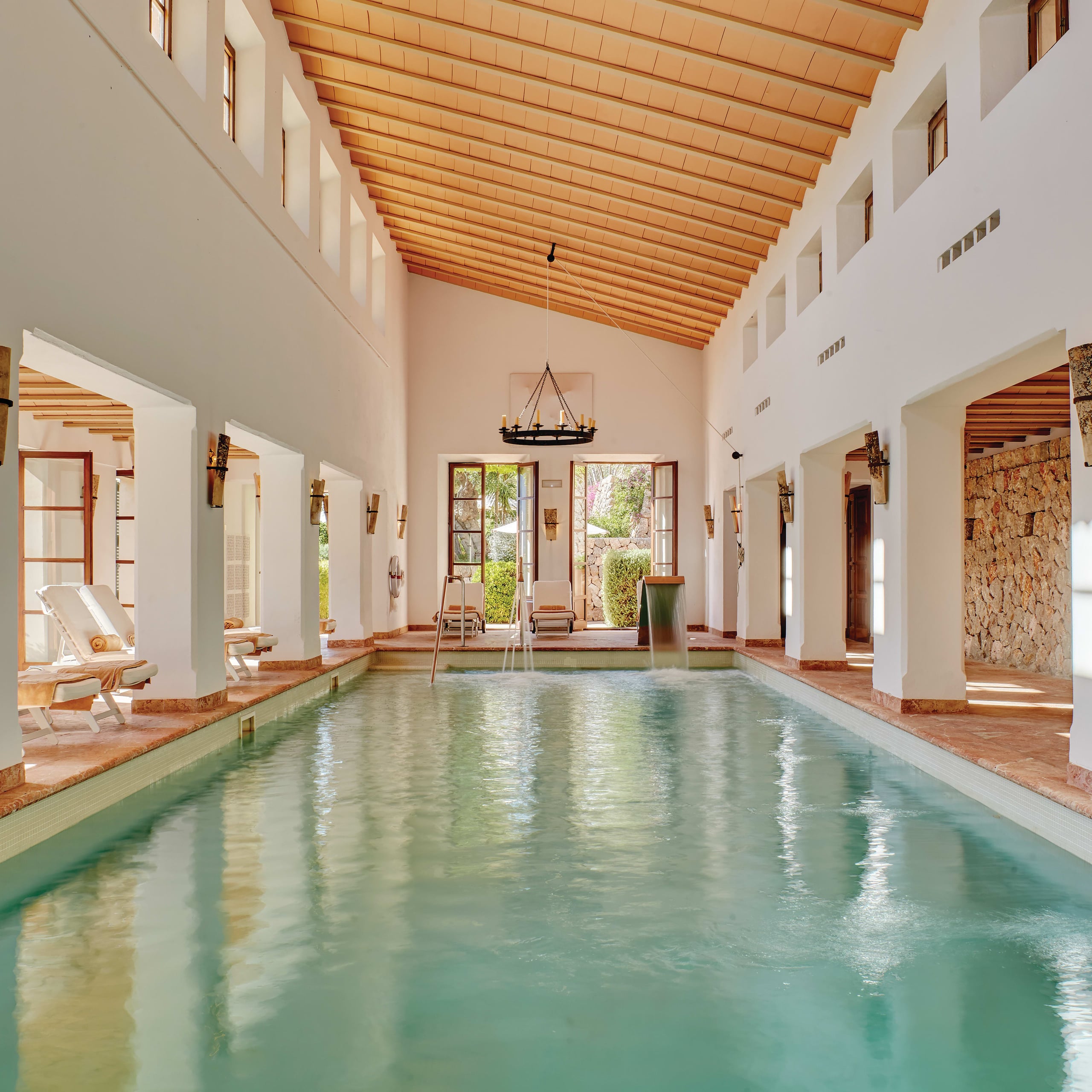 Belmond La Residencia, Balearic Islands, Spain — Indoor swimming pool with mineral-green water, exposed wooden beam ceiling, terracotta flooring, and arched colonnades opening to gardens.