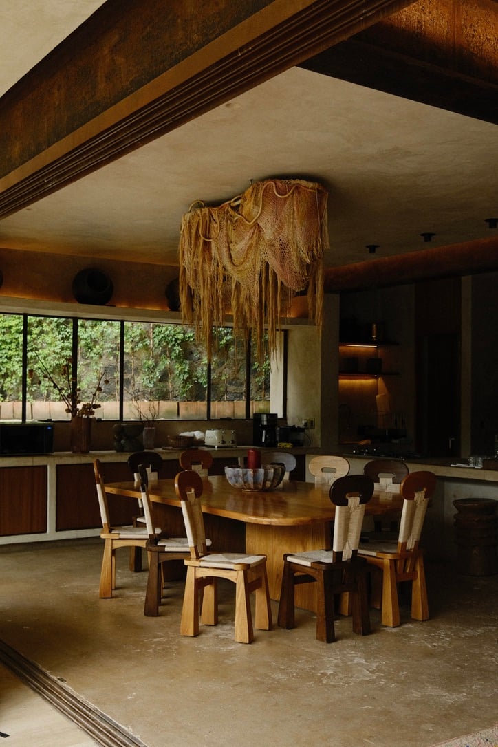 Casa Lumeria, Mexico — Wooden dining table with natural chairs beneath draped fiber pendant light, exposed timber beam ceiling, floor-to-ceiling windows overlooking verdant landscape.