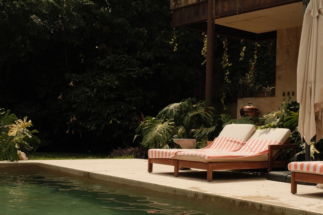 Casa Lumeria, Mexico — Wooden loungers with coral striped cushions beside green pool, lush tropical garden, modernist concrete pavilion with climbing vines.