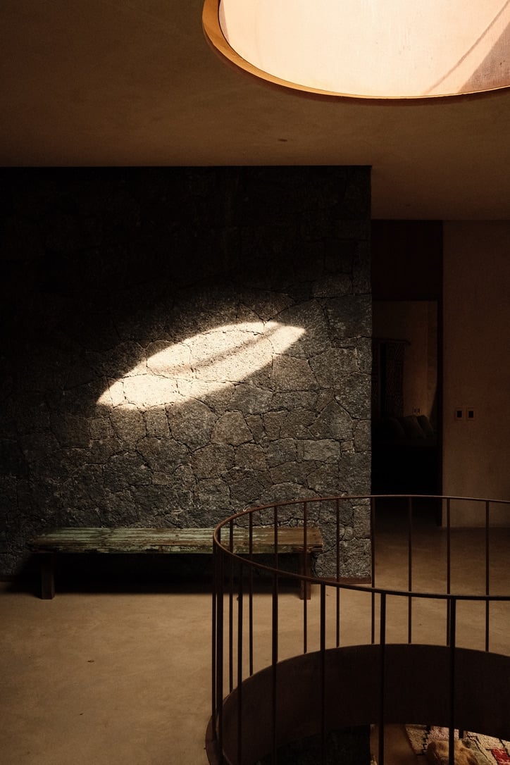 Casa Lumeria, Mexico — Brutalist stone gallery with curved metal railing, illuminated by dramatic oval skylight casting geometric light on textured wall.