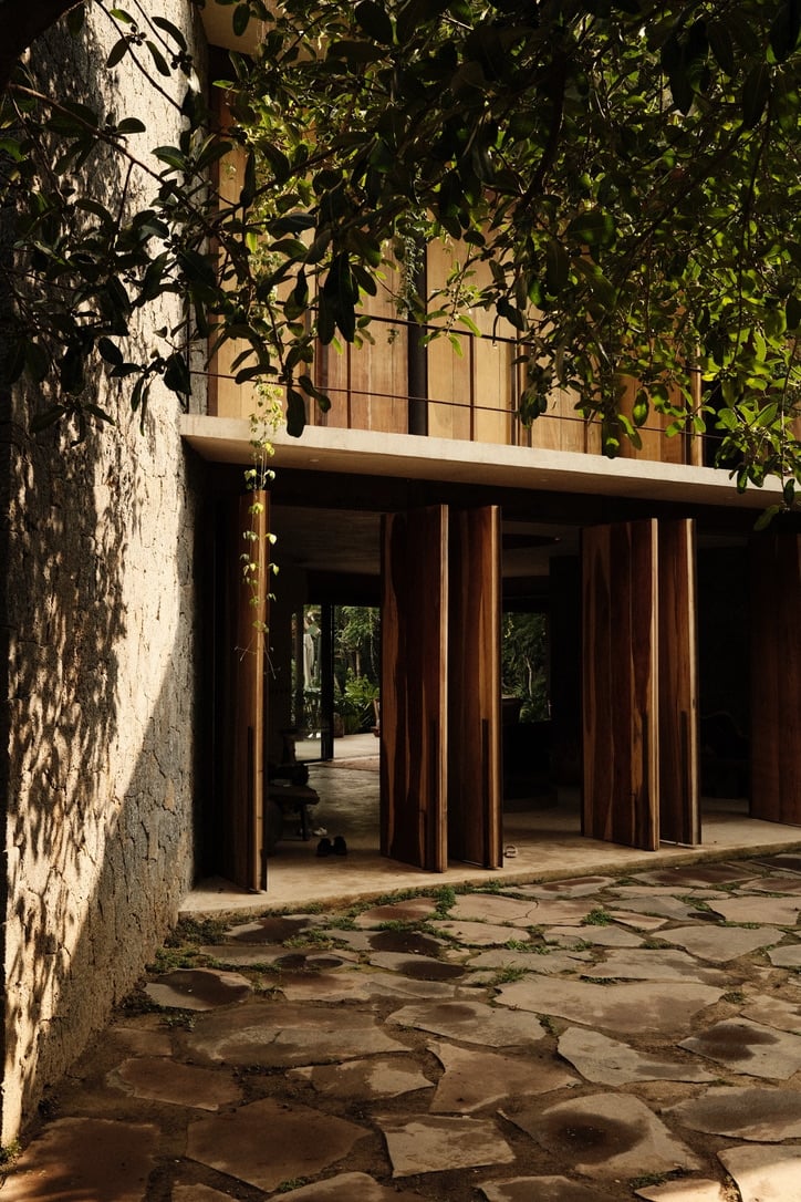 Casa Lumeria, Mexico — Modernist wooden colonnade opening onto stone courtyard, dappled sunlight through climbing vines, minimalist hospitality architecture.