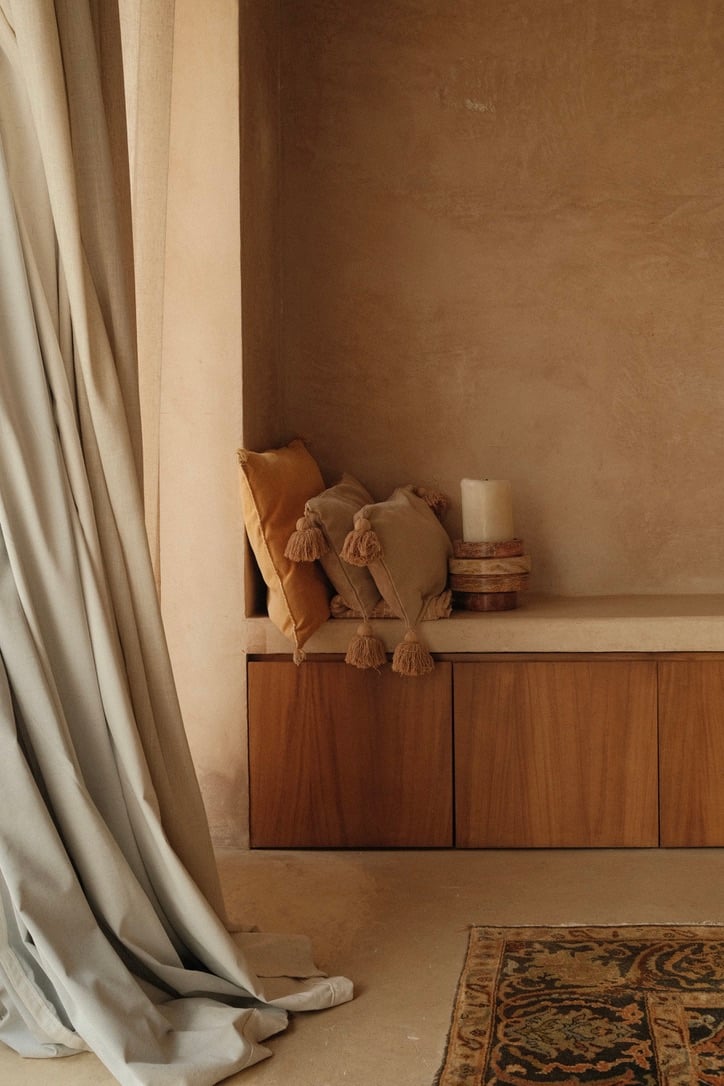 Casa Lumeria, Mexico — Minimalist bedroom alcove with warm plaster walls, wooden cabinetry, terracotta pillows with tassels, linen curtains, and patterned rug in natural light.