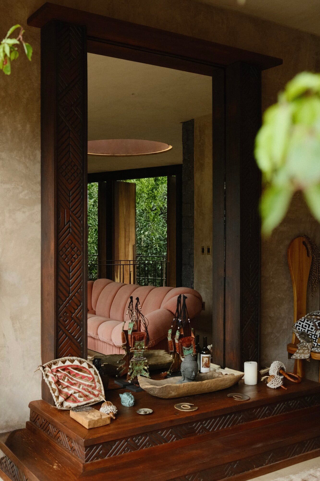 Casa Lumeria, Mexico — Dark wooden mirror frame with carved geometric detailing above console displaying artisan ceramics, leather accessories, and woven textiles, reflecting pink curved sofa and verdant garden beyond.