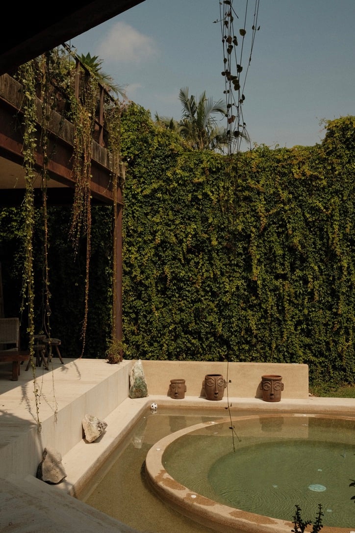 Casa Lumeria, Mexico — Sunlit courtyard with curved plunge pool, terracotta sculptural heads, cream stone surround, and dense ivy-covered walls creating serene tropical retreat.