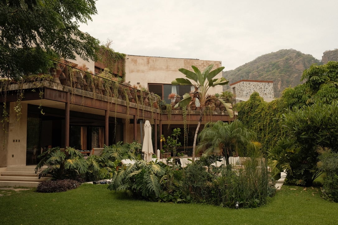 Casa Lumeria, Mexico — Modernist villa with weathered terracotta façade, lush tropical garden, mountain backdrop, covered terrace with white umbrellas and native plantings.