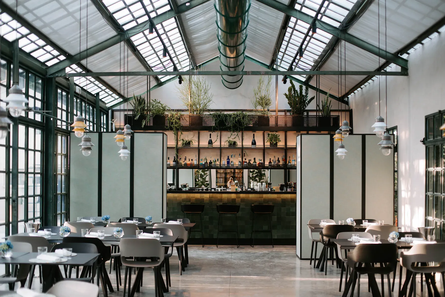 Ristorante Limonaia, Europe — Minimalist dining room with green steel framework, skylight ceiling, suspended pendant lights, and bar with living plant wall backdrop.