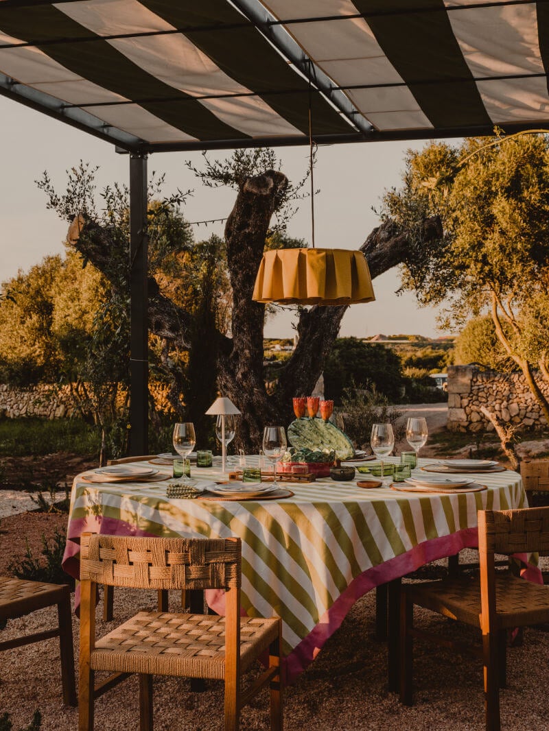 Amagatay Restaurant, Balearic Islands, Spain — Outdoor dining under retractable pergola with striped tablecloth, vintage pendant lamp, gnarled olive trees, golden hour light.