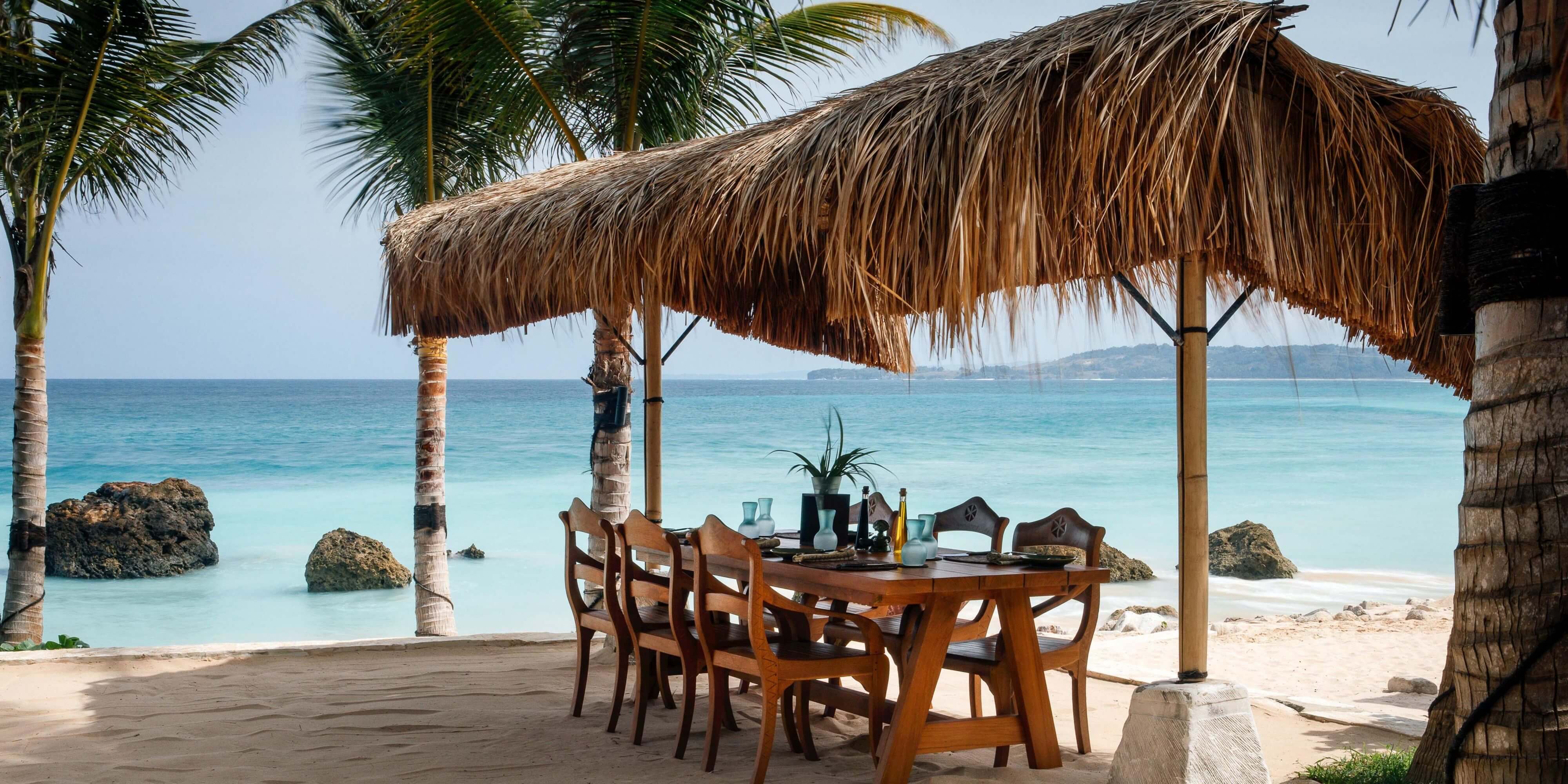 NIHI Sumba, Asia — Thatched palapa dining pavilion with wooden table and chairs overlooking turquoise ocean, white sand beach, and rocky outcrops at sunset.
