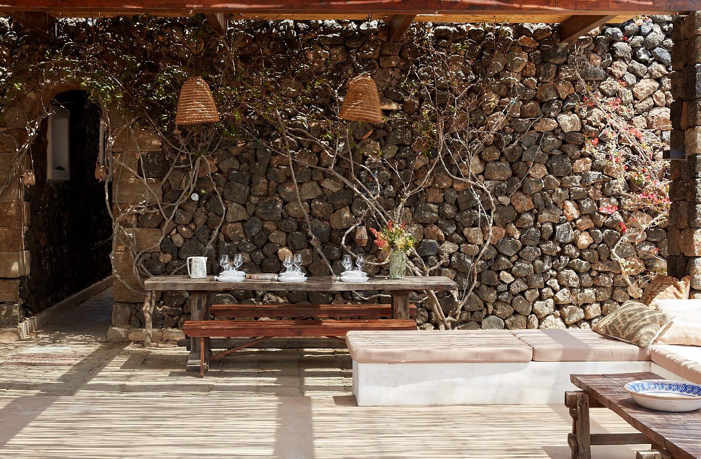 Parco Dei Sesi, Europe — Stone terrace with woven pendant lights, weathered wooden dining table, linen seating, climbing vines, and Mediterranean architecture in dappled sunlight.