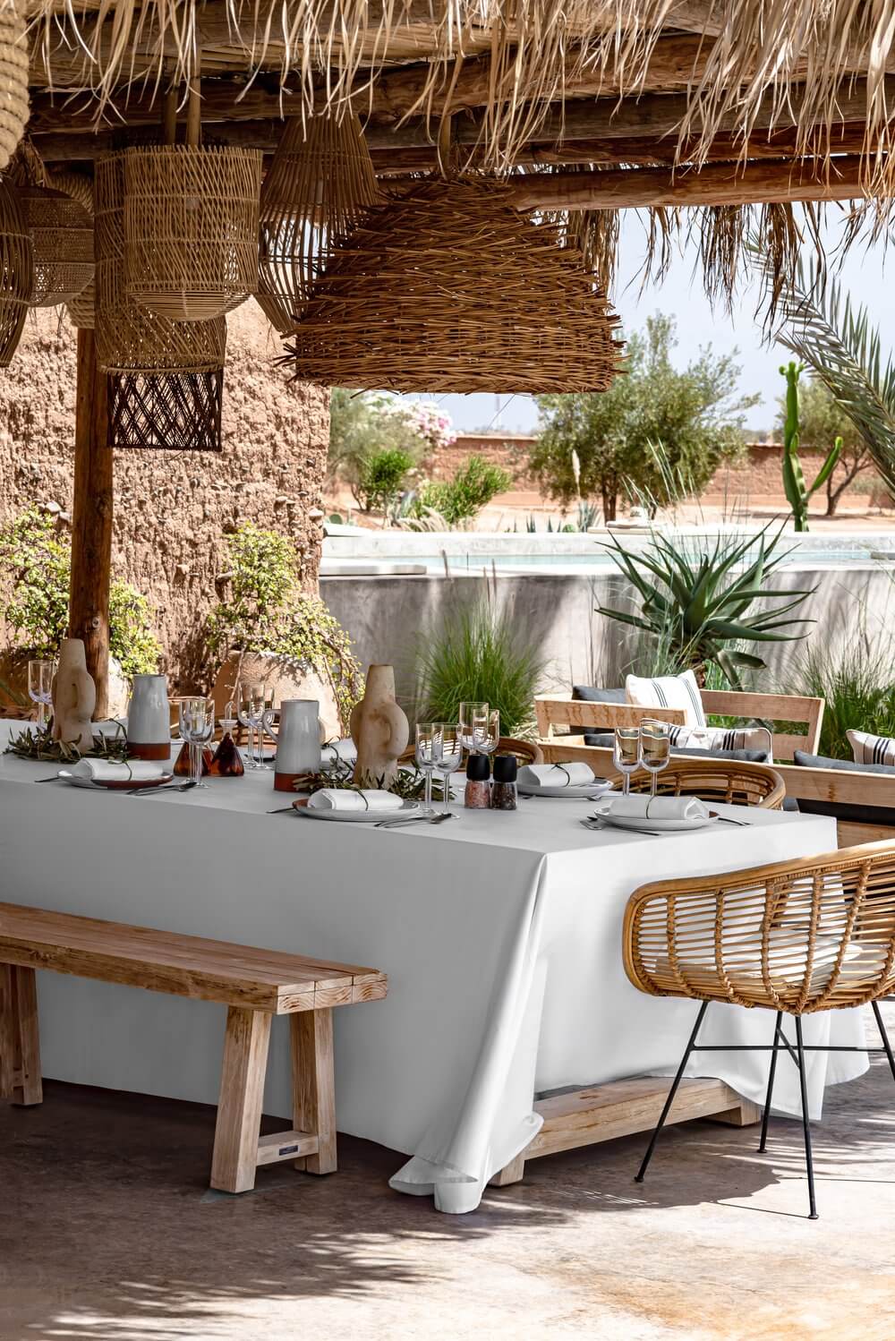 The Kitchen, Africa — Outdoor dining terrace with woven palm pergola, white linen-draped table, rattan chairs, desert landscape, and clay vessel accents.