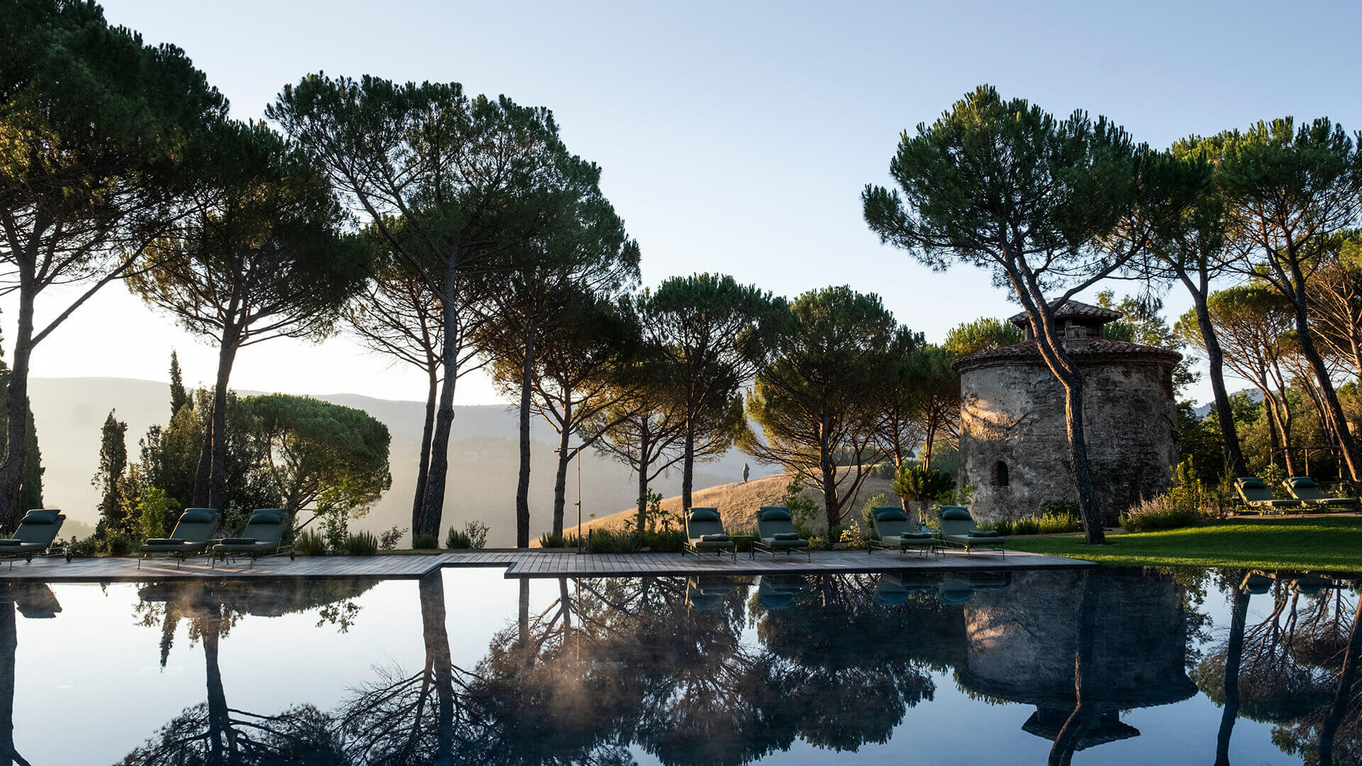 Castello di Reschio, Tuscany, Italy — Infinity pool with stone tower and Italian stone pines reflecting at sunset, minimalist loungers overlooking rolling hills.