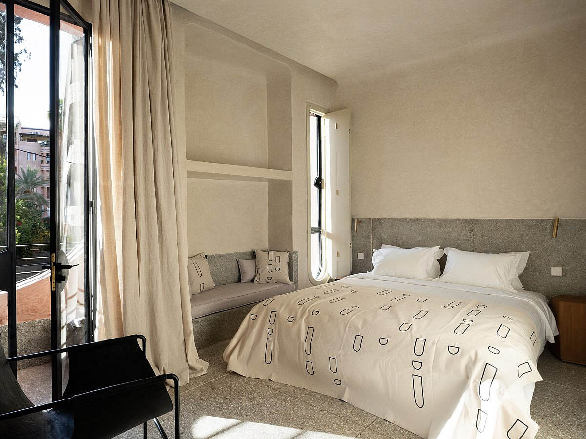 Maison Brummell Majorelle, Africa — Minimalist bedroom with taupe walls, linen bedding, gray upholstered headboard, alcove seating, and French doors opening to terrace light.