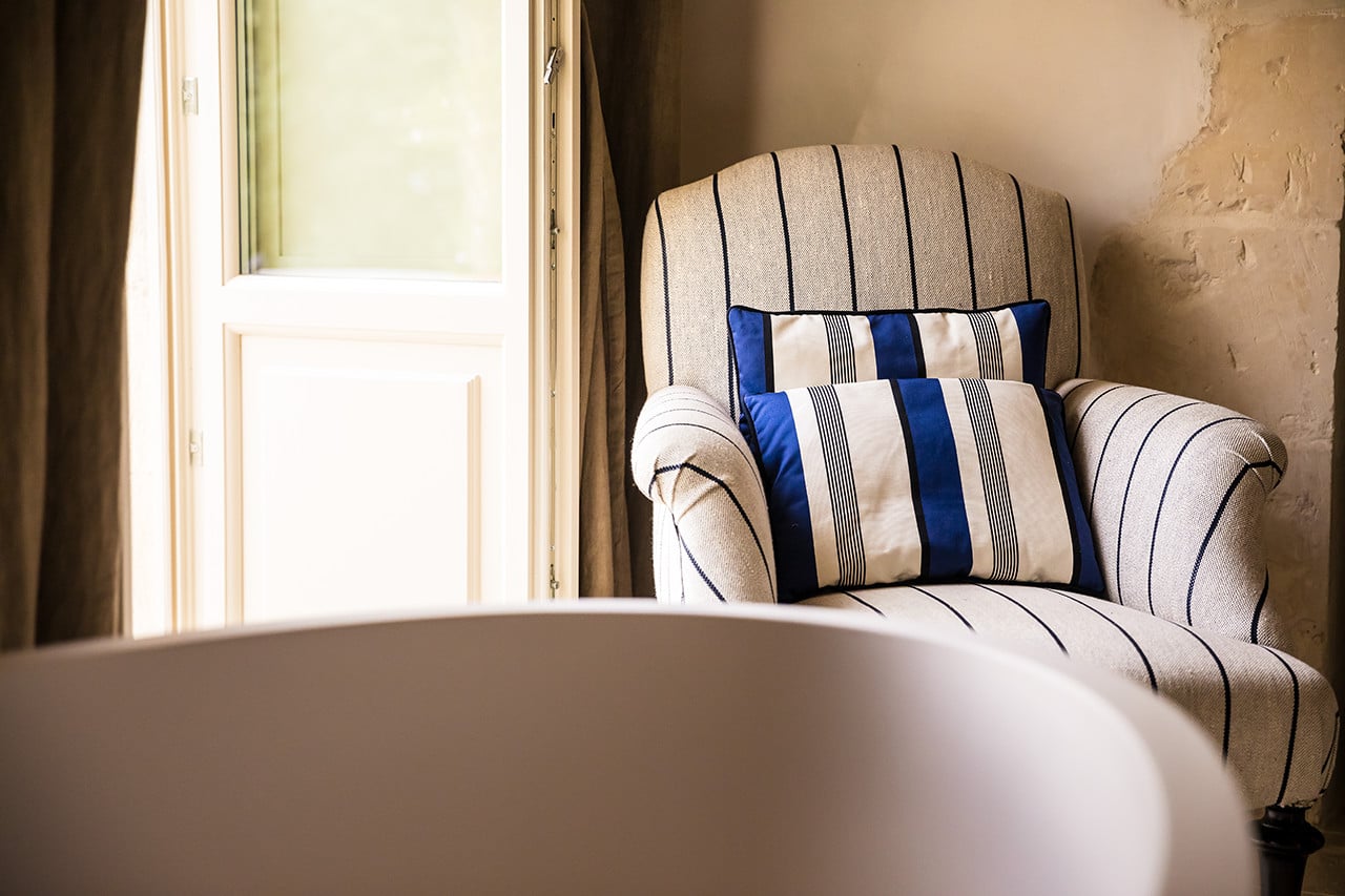 Dimora delle Balze, Europe — Striped armchair with navy and cream pillows beside window with natural light and textured plaster walls.