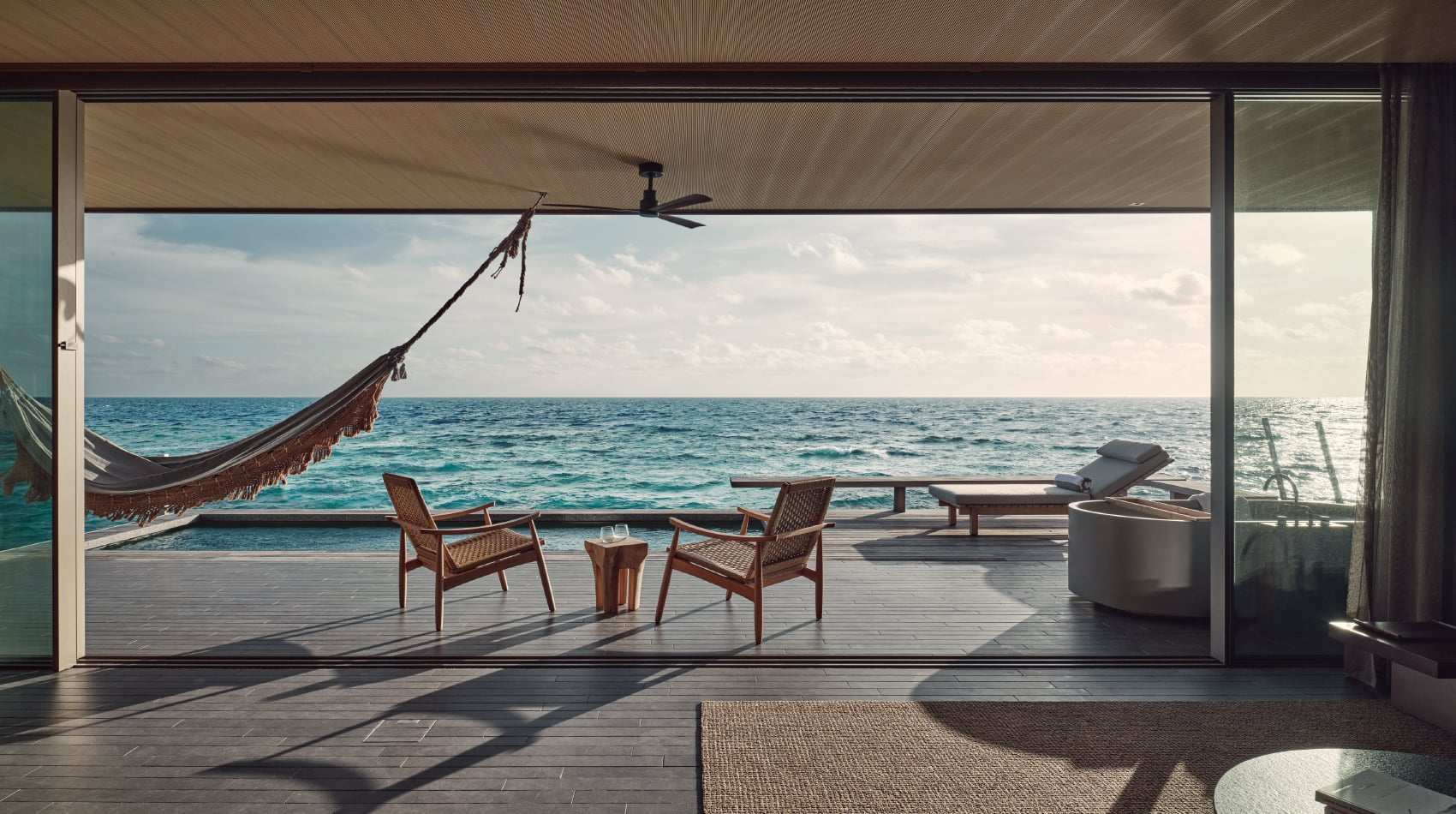Patina Maldives, Asia — Overwater villa deck with teak lounge chairs, ocean view, hanging hammock, ceiling fan, and expansive glass doors opening to turquoise sea.
