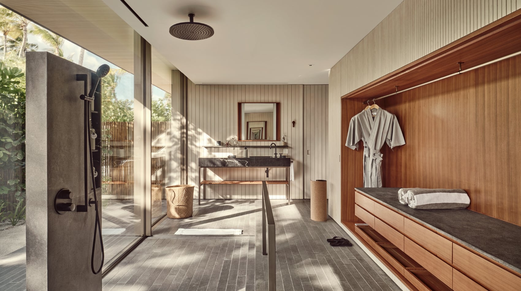 Patina Maldives, Asia — Spa bathroom with teak cabinetry, concrete shower, terrazzo sink, rainfall showerhead, and garden views through floor-to-ceiling glass.