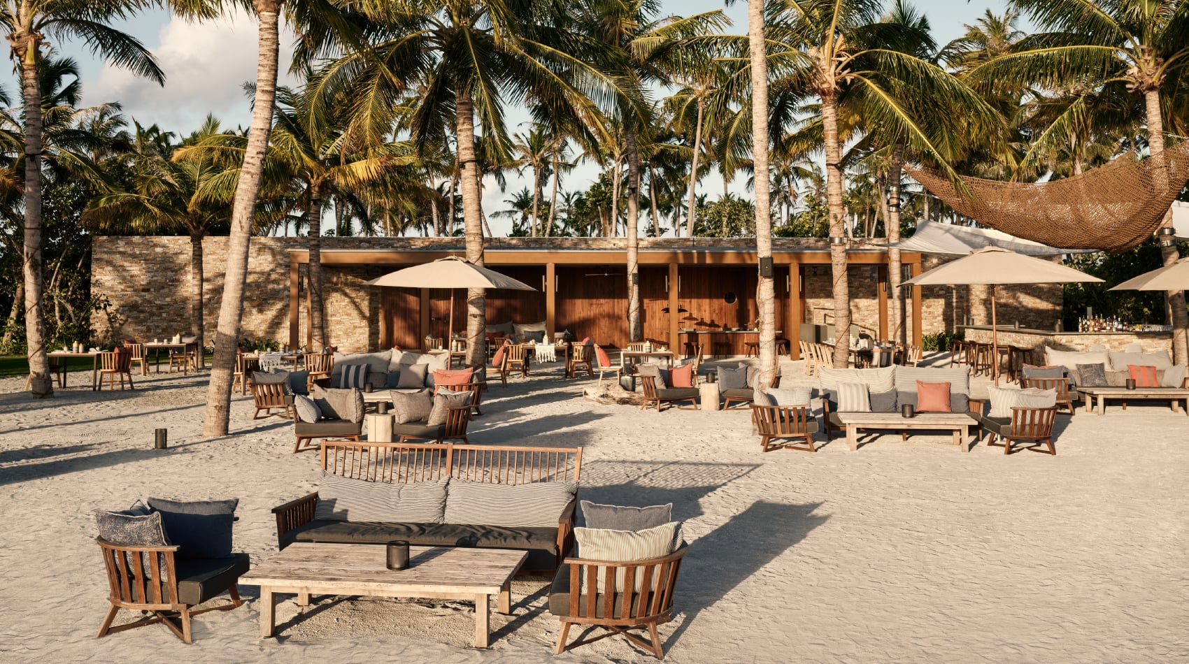 Helios, Asia — Beach pavilion with wooden furniture, palm trees, stone architecture, and cream umbrellas creating a serene luxury resort setting.