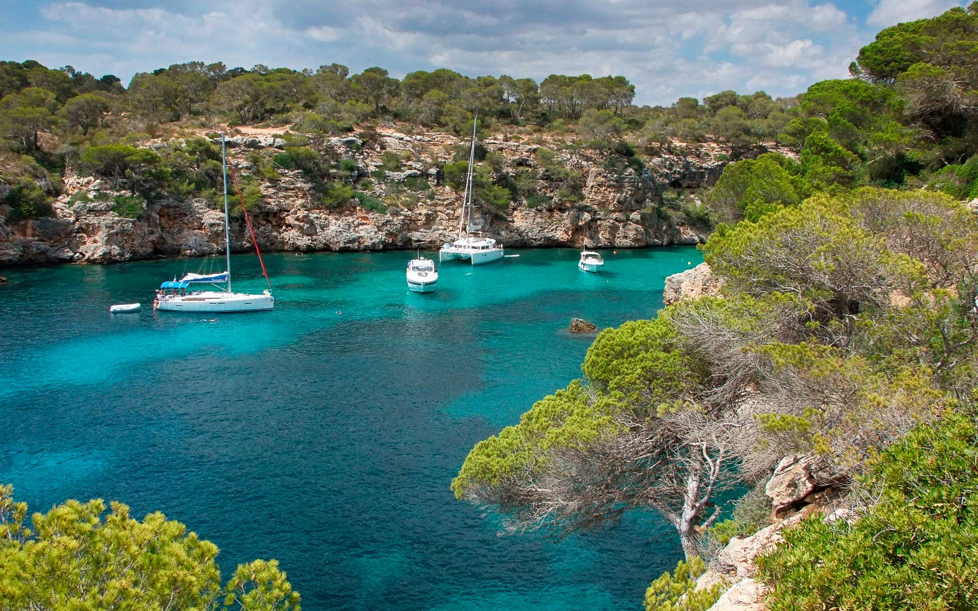 Sailing Day, Balearic Islands, Spain — Moored sailboats in turquoise Mediterranean cove surrounded by limestone cliffs and pine forest.