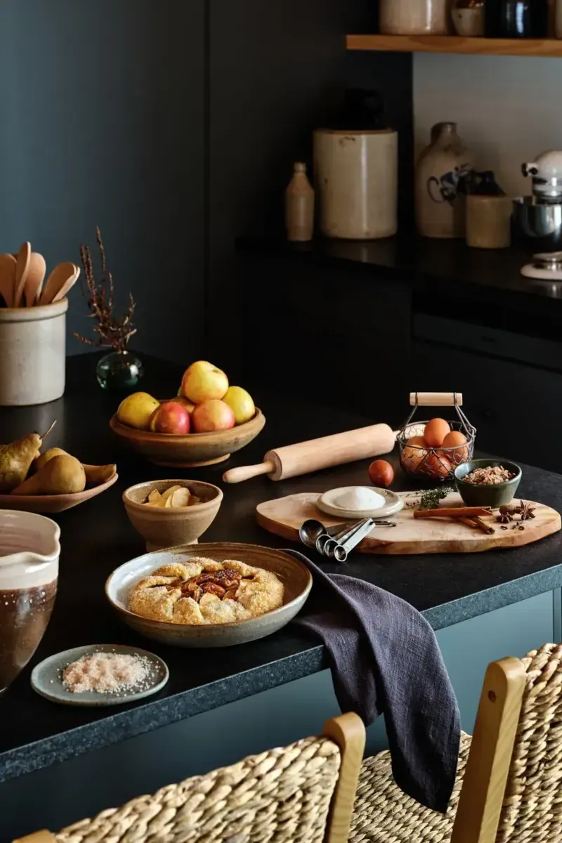 Wildflower Farms, North America — Black kitchen counter styled with fresh apples, baked galette, wooden utensils, copper eggs basket, and neutral pottery against dark walls and teal cabinetry.