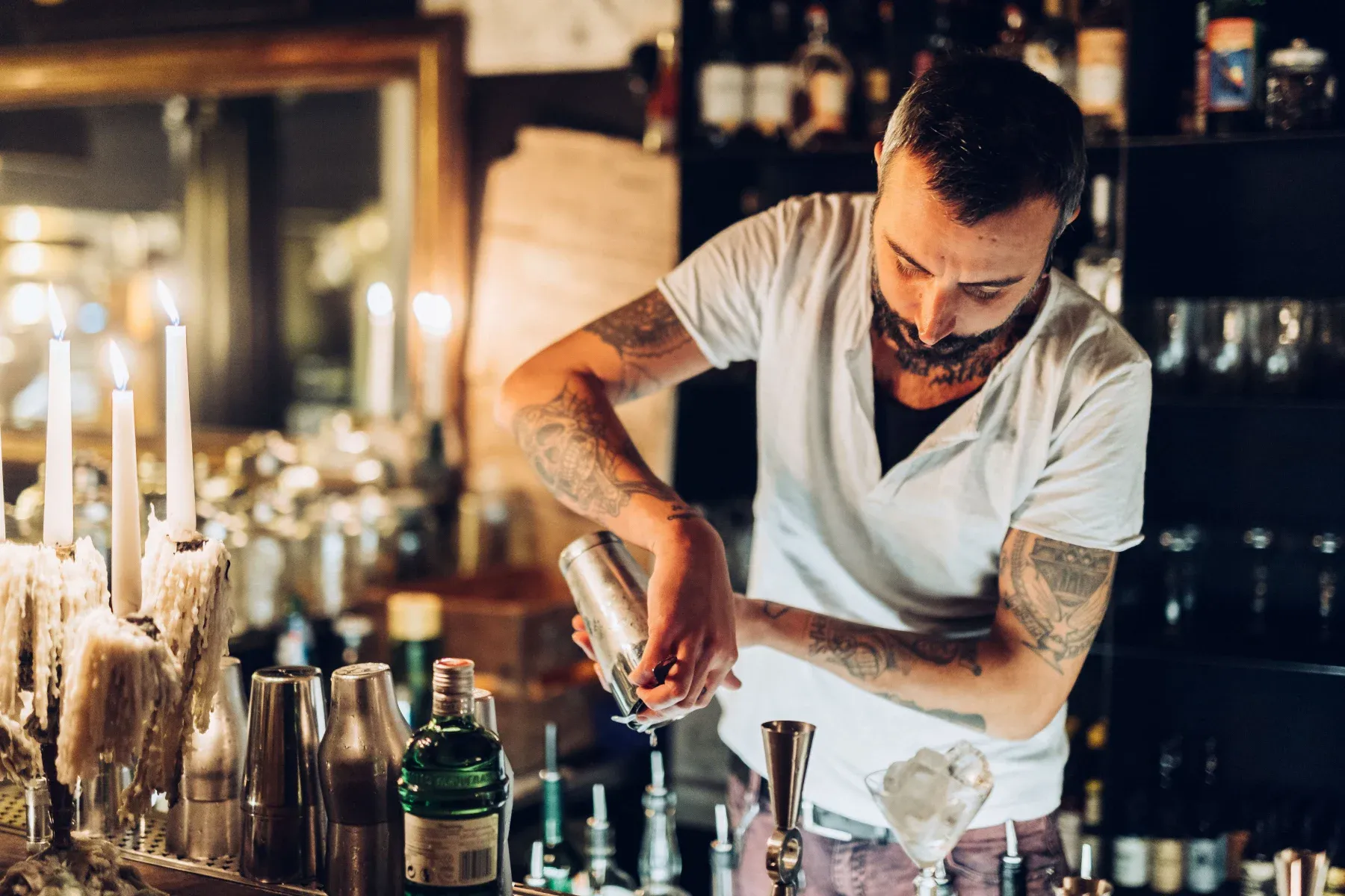 Herbarium Bar, Europe — Bartender crafting cocktail at candlelit counter with brass shakers and glassware, intimate evening ambiance.
