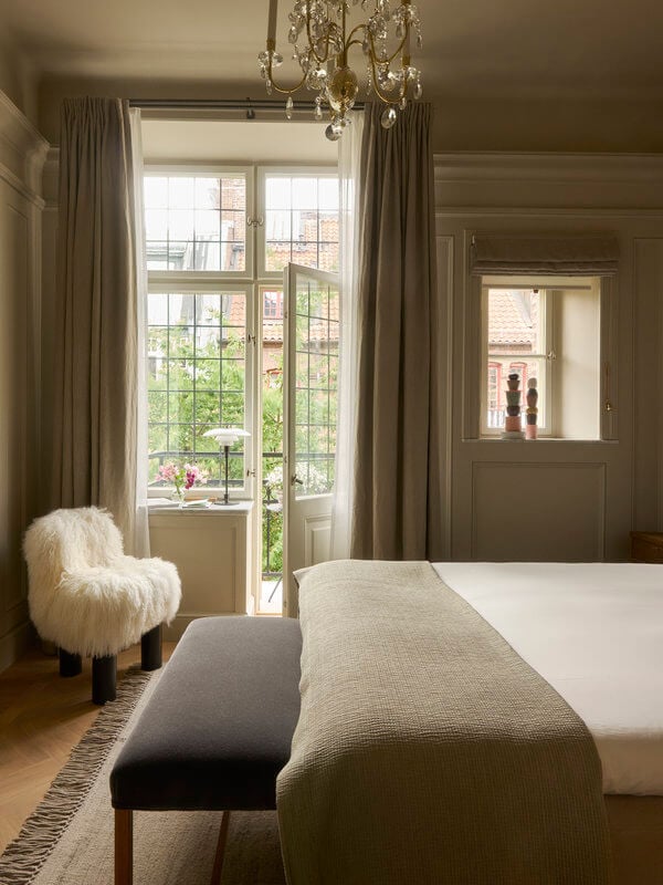 Ett Hem, Europe — Serene bedroom with French doors opening to garden, cream linen bedding, crystal chandelier, taupe walls, and white fur chair in natural light.
