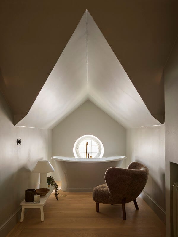 Ett Hem, Europe — Minimalist spa sanctuary with peaked white ceiling, soaking tub, glowing circular mirror, brown curved chair, and warm wood flooring under soft architectural light.