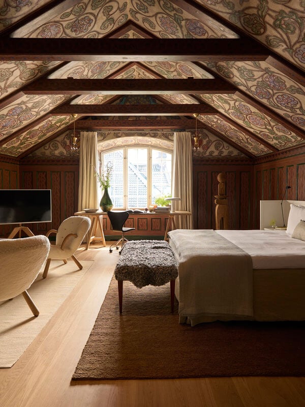 Ett Hem, Stockholm, Sweden — Vaulted attic bedroom with exposed beams, patterned ceiling panels, wood paneling, mid-century chairs, and natural light from dormer window.