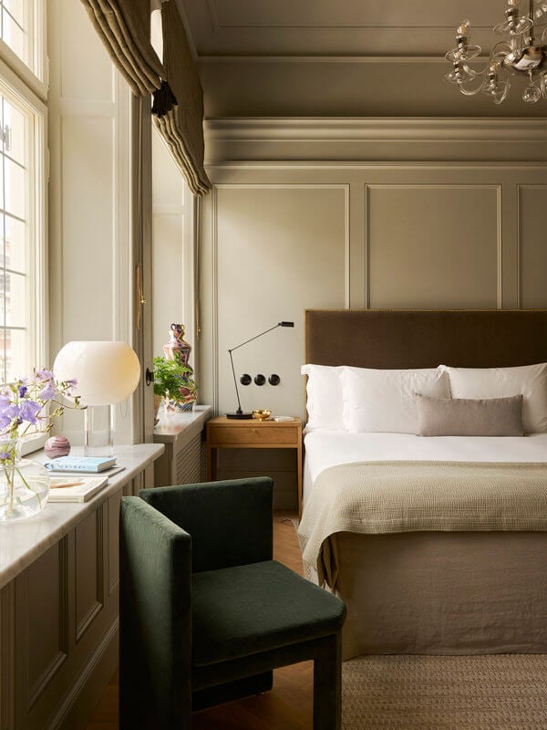 Ett Hem, Europe — Sunlit bedroom with taupe paneled walls, brown upholstered headboard, white bedding, green accent chair, wooden nightstand, and crystal chandelier.
