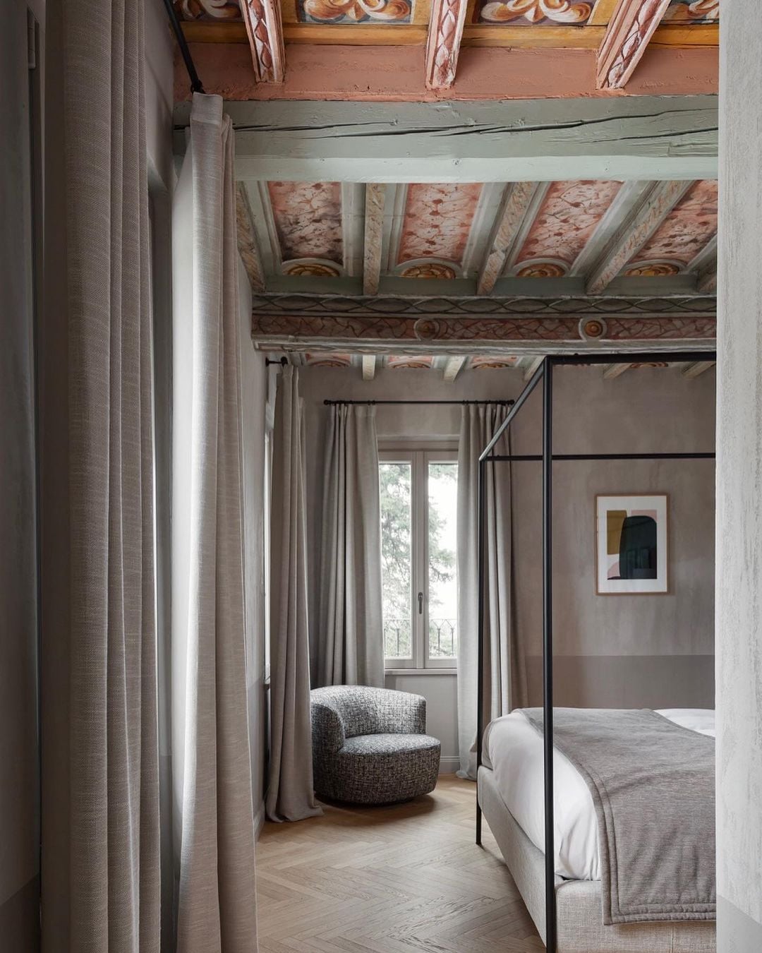 Roncolo 1888, Europe — Bedroom with exposed timber ceiling beams, frescoed details, canopy bed, curved armchair, and floor-to-ceiling curtains overlooking landscape.