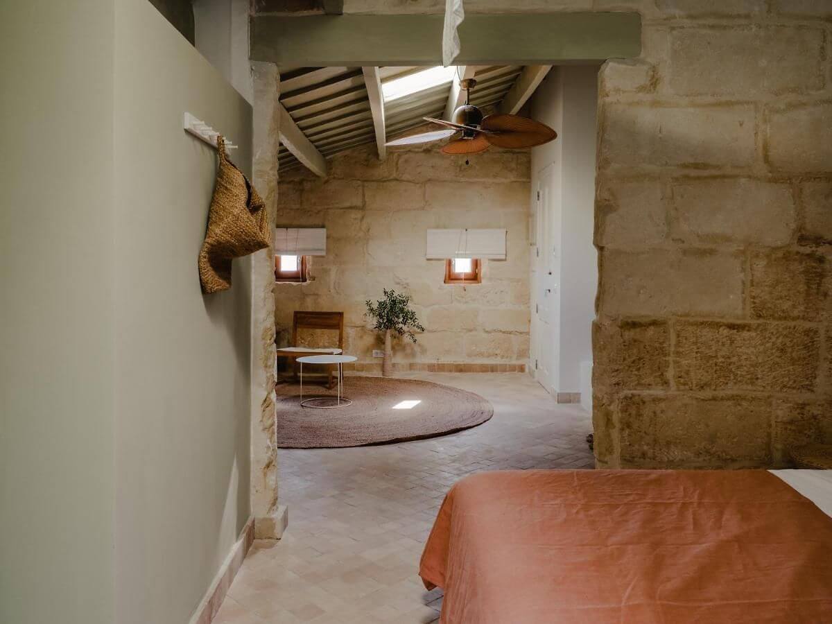 Amagatay, Balearic Islands, Spain — Stone-walled studio with exposed wooden beams, ceiling fan, terracotta floor, and natural light from high windows.