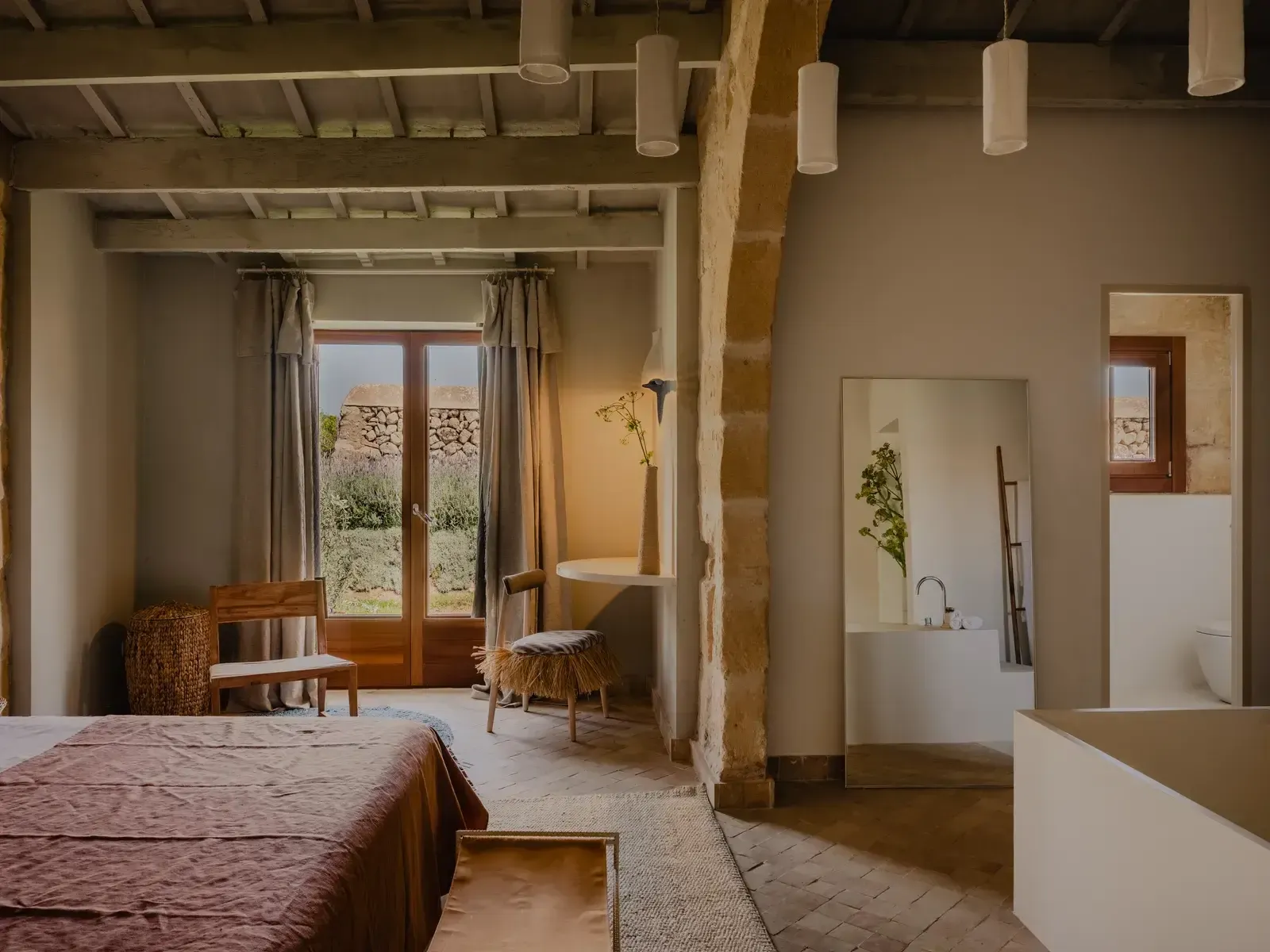 Amagatay, Balearic Islands — Minimalist bedroom with exposed stone columns, terracotta floors, wooden furnishings, pendant lights, and garden views through open doors.