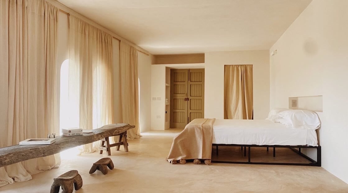 Parco Dei Sesi, Europe — Minimalist bedroom with natural linen curtains, driftwood sculpture, wooden bed frame, and warm stone flooring bathed in soft daylight.