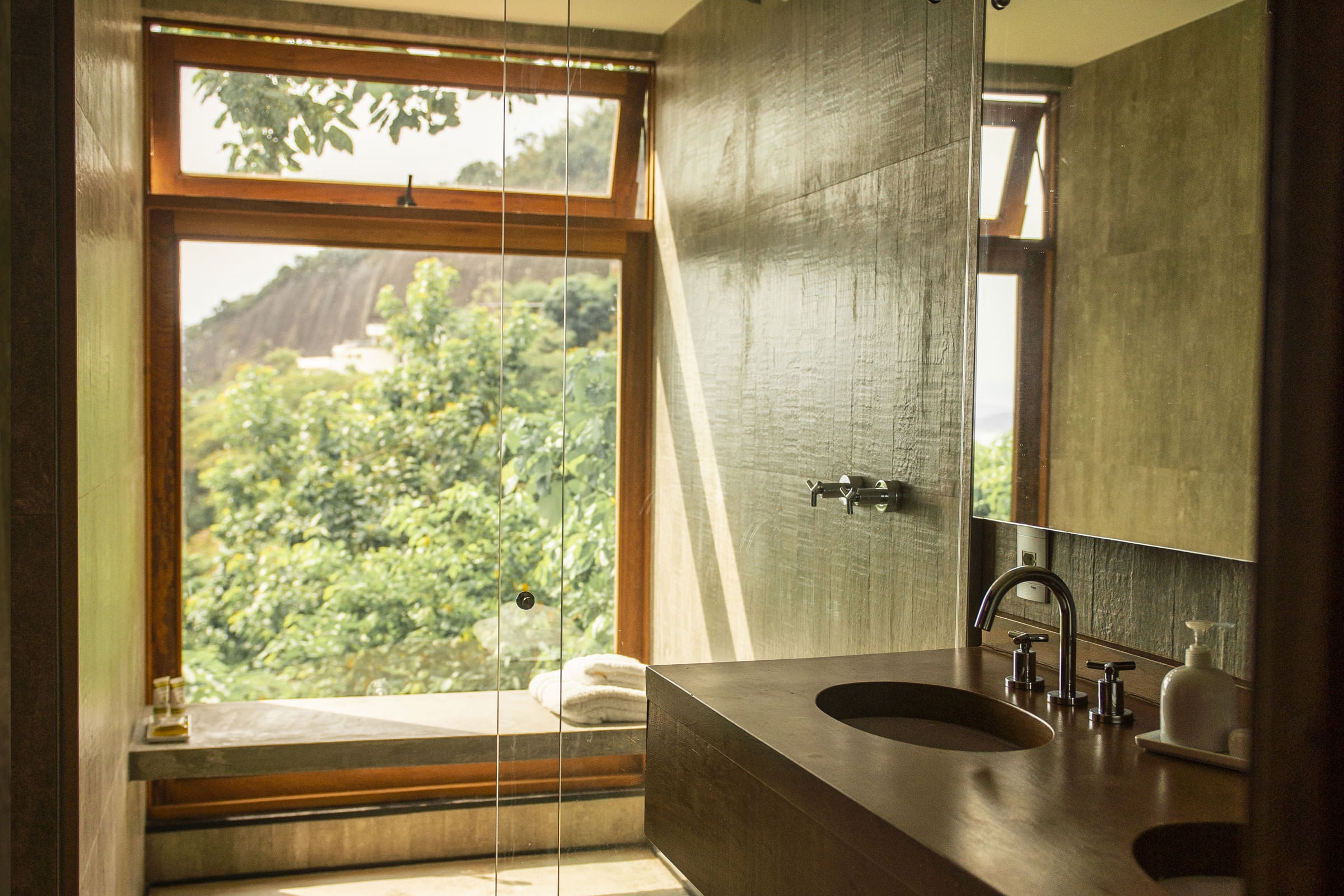 Chez Georges, South America — Minimalist bathroom with dark stone sink, floor-to-ceiling mirror, wooden-framed windows overlooking lush hillside landscape.