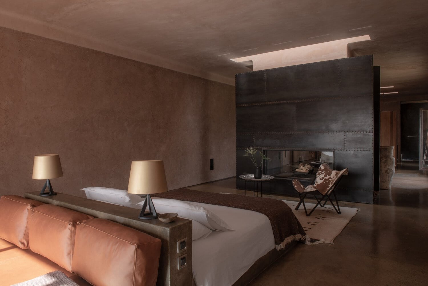 Villa D, Africa — Modernist bedroom with textured terracotta walls, linear ceiling light, paired beds with brass lamps, black steel fireplace feature, and leather seating in warm earthy tones.