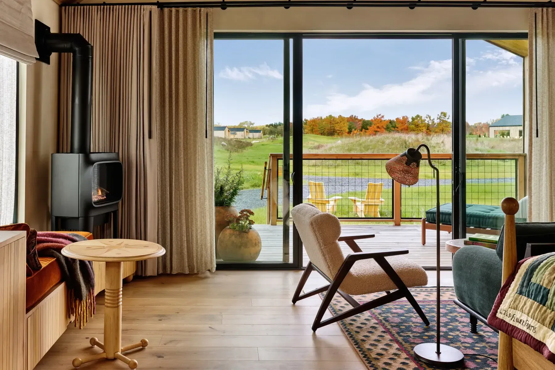 Wildflower Farms, North America — Modern villa interior with wood stove, mid-century chair, and floor-to-ceiling windows overlooking pastoral landscape with autumn trees.