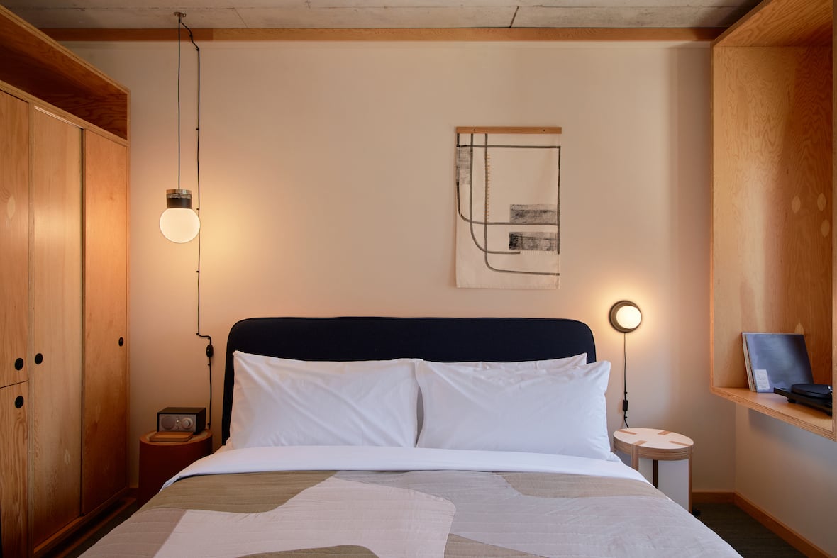 Ace Hotel Toronto — Minimalist bedroom with black headboard, white linens, warm wood cabinetry, pendant lighting, and abstract wall art in neutral tones.