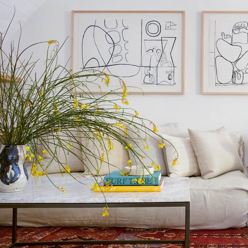 The Surfrider, North America — Minimalist living room with line-art framed prints, cream linen sofa, dried grass arrangement, and design books on marble coffee table.