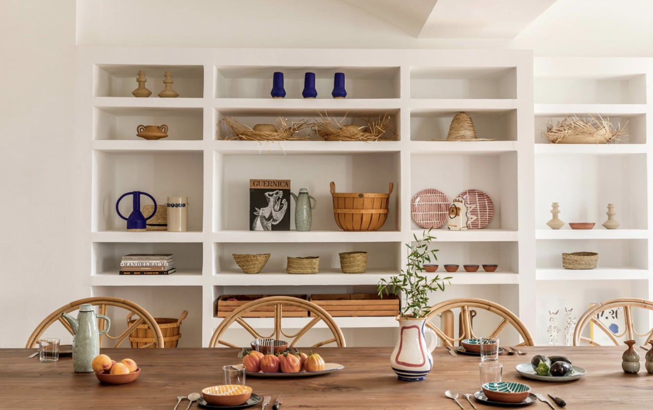 Menorca Experimental, Balearic Islands — Dining room with built-in shelving, natural wood table, woven chairs, artisan ceramics, and curated home goods displaying Mediterranean design aesthetic.