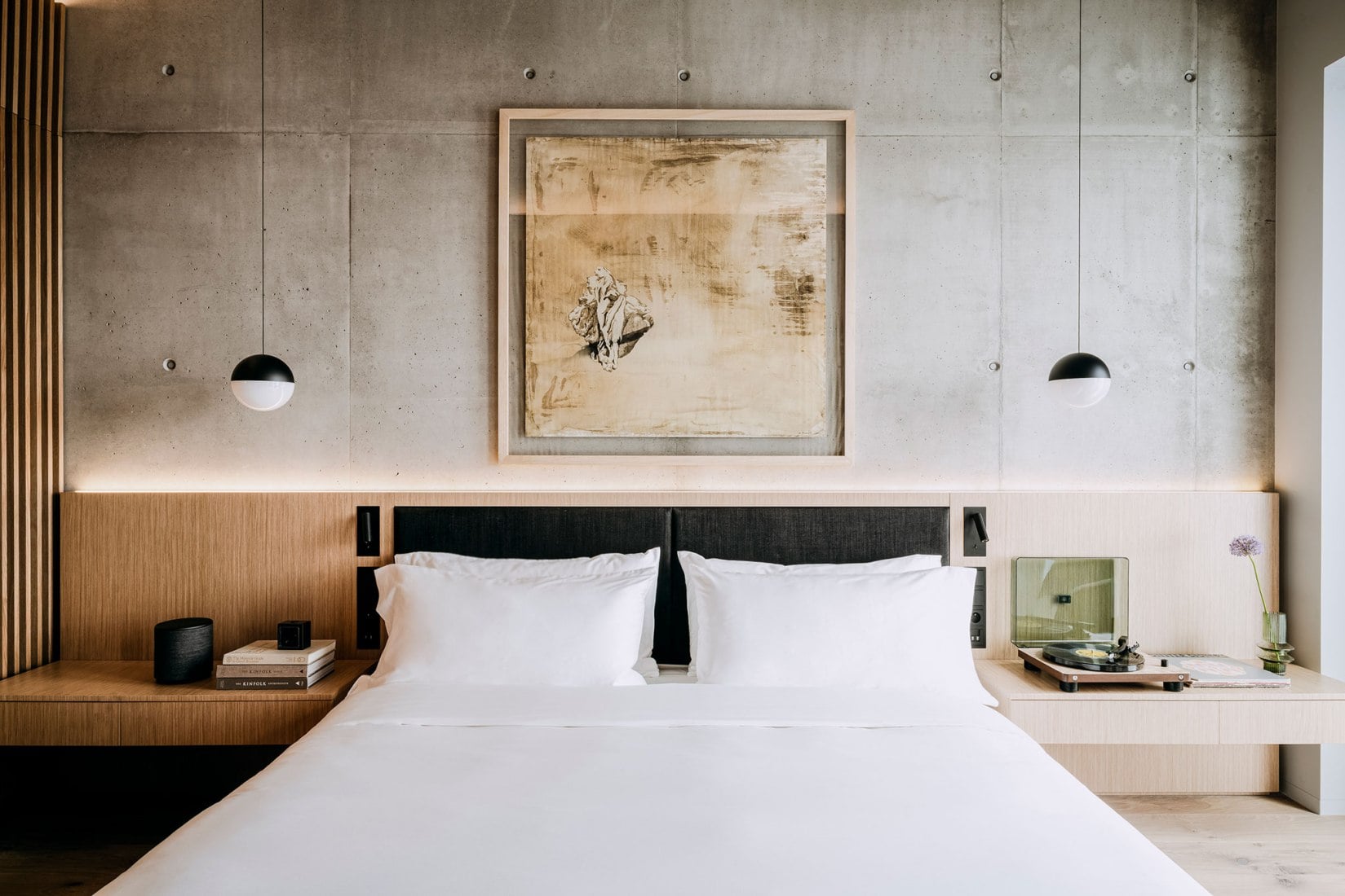 Nobu Warsaw, Poland — Minimalist bedroom with concrete walls, wood-paneled bedside tables, framed botanical artwork, pendant lighting, and crisp white linens.