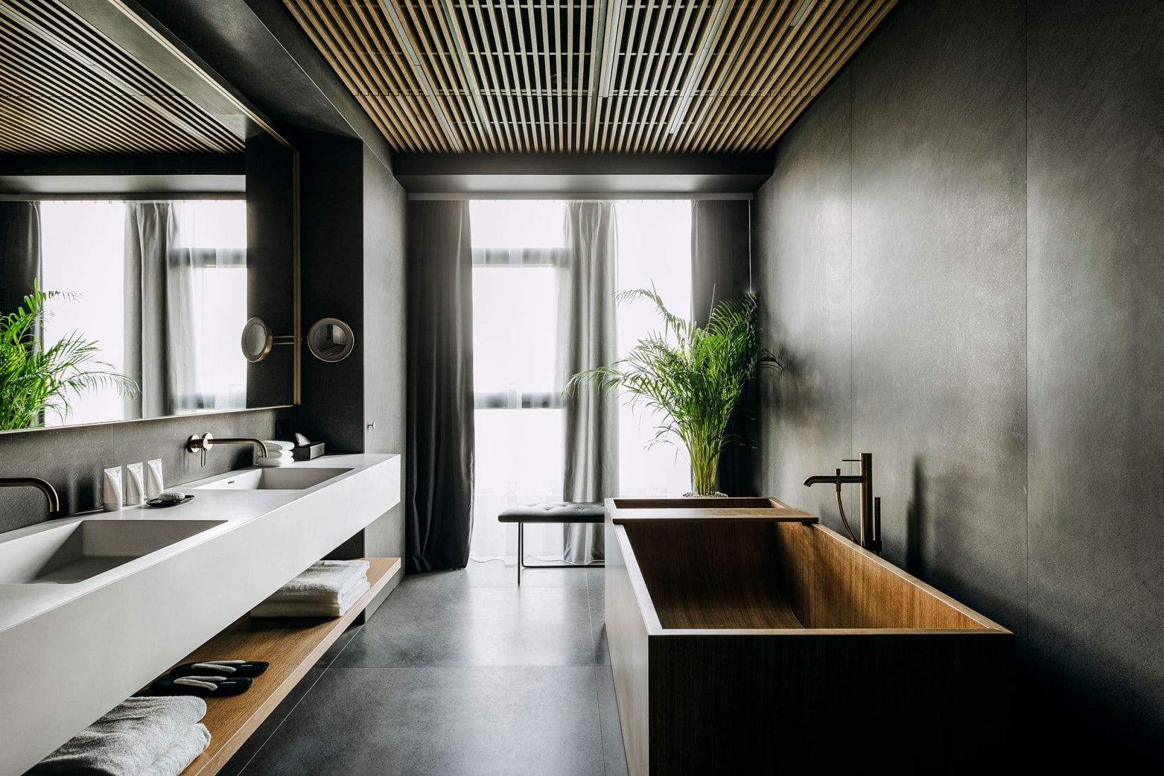 Nobu Warsaw, Poland — Minimalist spa bathroom with black walls, wooden soaking tub, double vanity, slatted ceiling, and floor-to-ceiling windows with city views.
