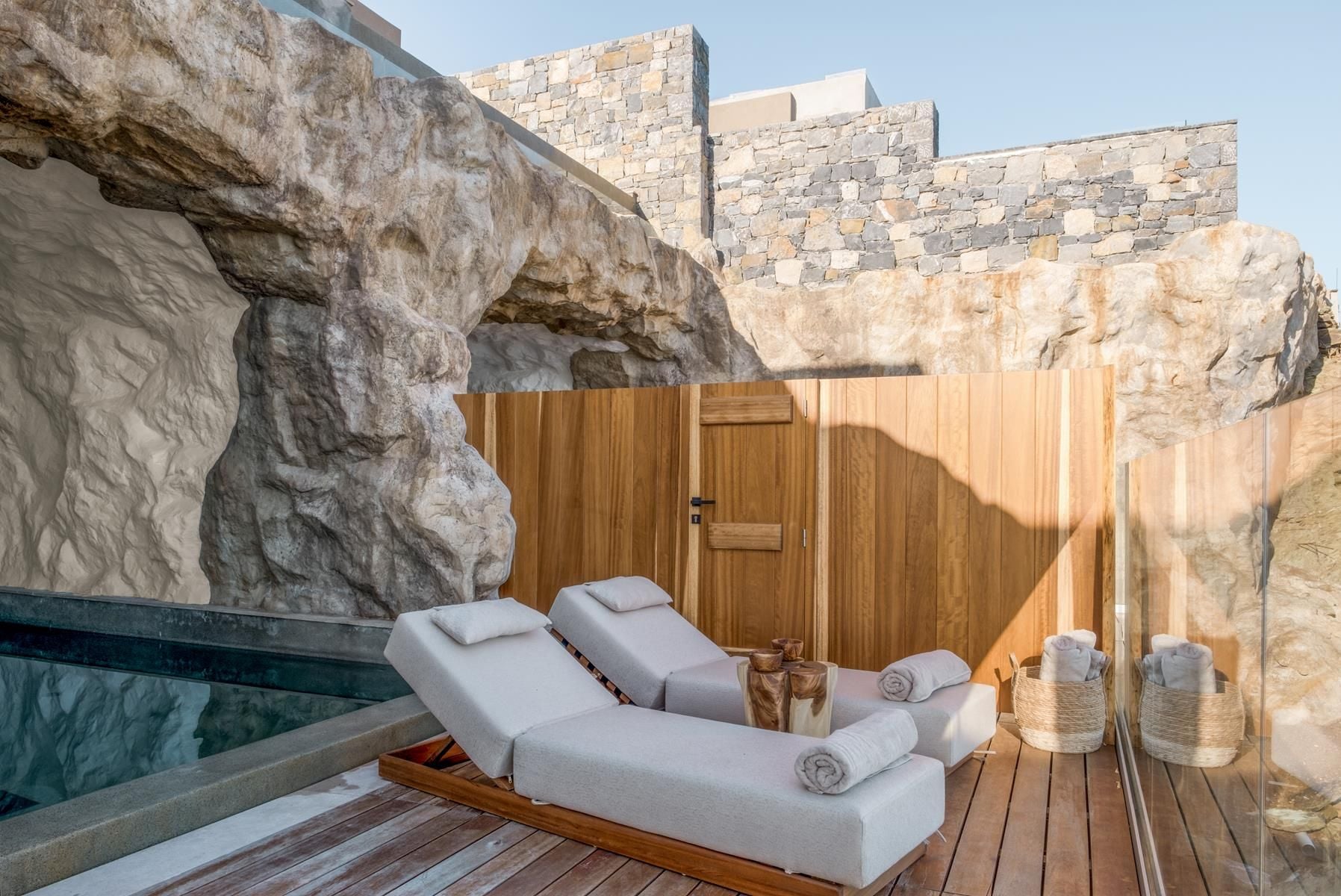 ACRO Suites, Europe — Limestone spa terrace with three loungers, wooden screen, rolled towels, and natural stone walls beneath clear sky.