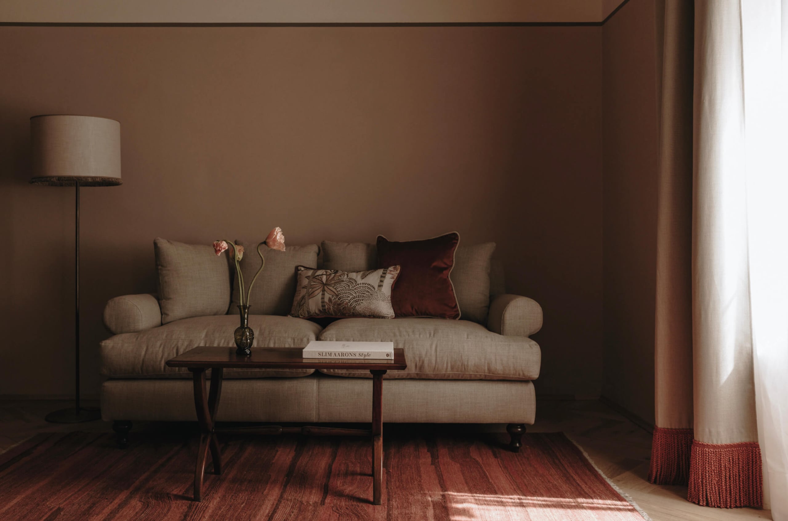Parkhotel Mondschein, Europe — Beige linen sofa with decorative pillows, dark wood side table, floor lamp, warm terracotta walls, natural light from draped curtains, minimalist luxury interior.