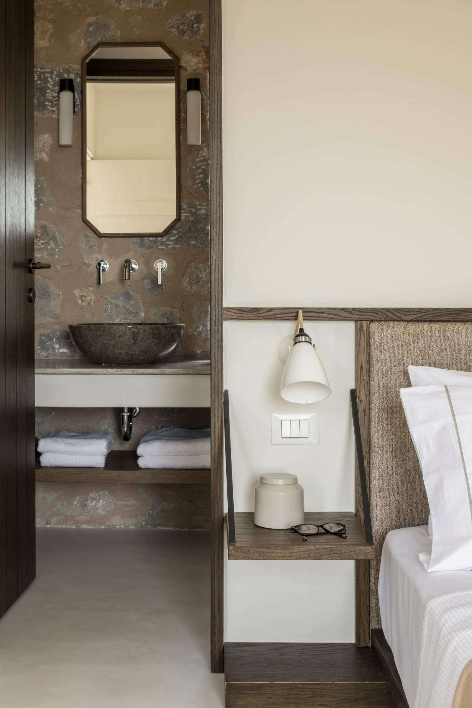 Monolithoi, Europe — Ensuite bathroom with stone vessel sink, framed mirror, wall sconces, and adjoining bedroom with natural wood nightstand and pendant lighting.