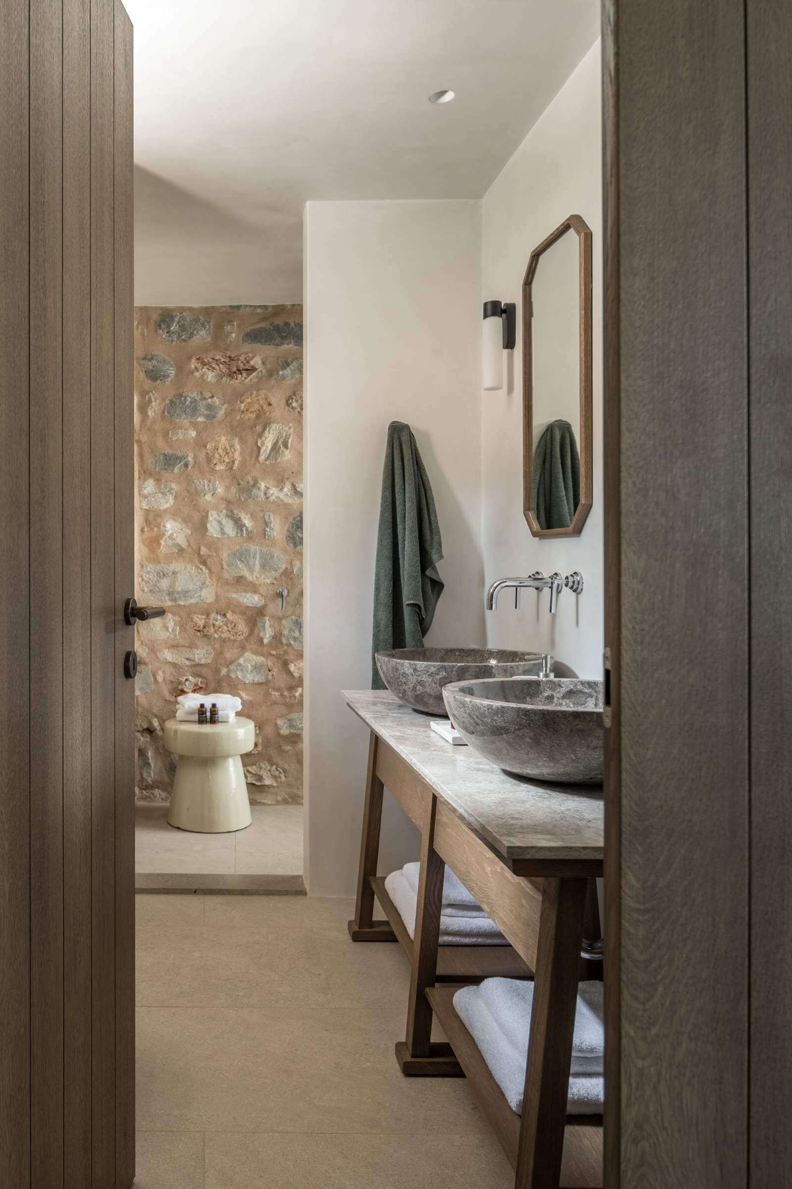 Monolithoi, Europe — Spa bathroom with dual stone vessel sinks, wooden vanity, arched mirror, natural stone accent wall, soft green linens, minimalist luxury design.