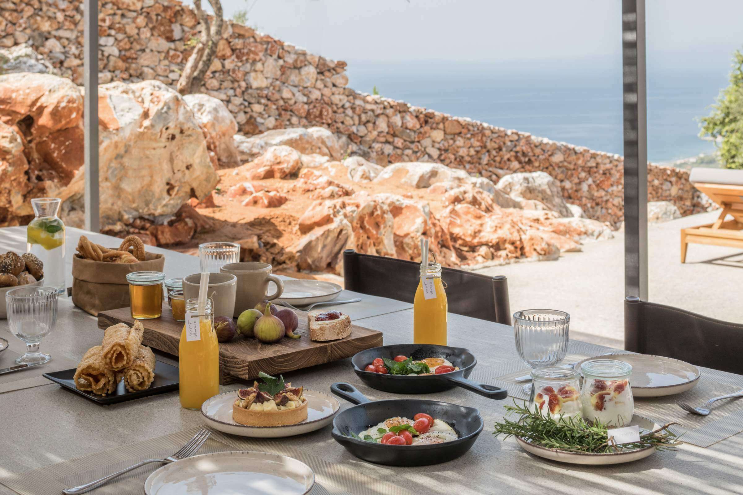 Monolithoi, Europe — Clifftop dining table with Mediterranean salads, citrus beverages, and stone boulder landscape views through floor-to-ceiling windows.