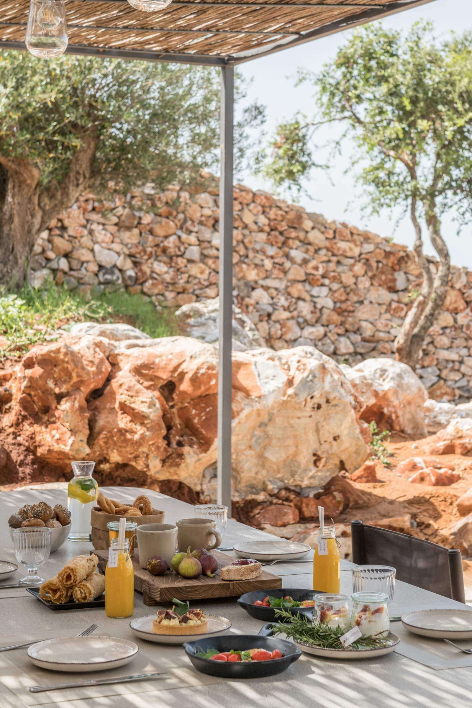 Monolithoi, Europe — Outdoor dining under pergola with stone walls, olive trees, fresh Mediterranean breakfast spread, natural light and rustic architecture.