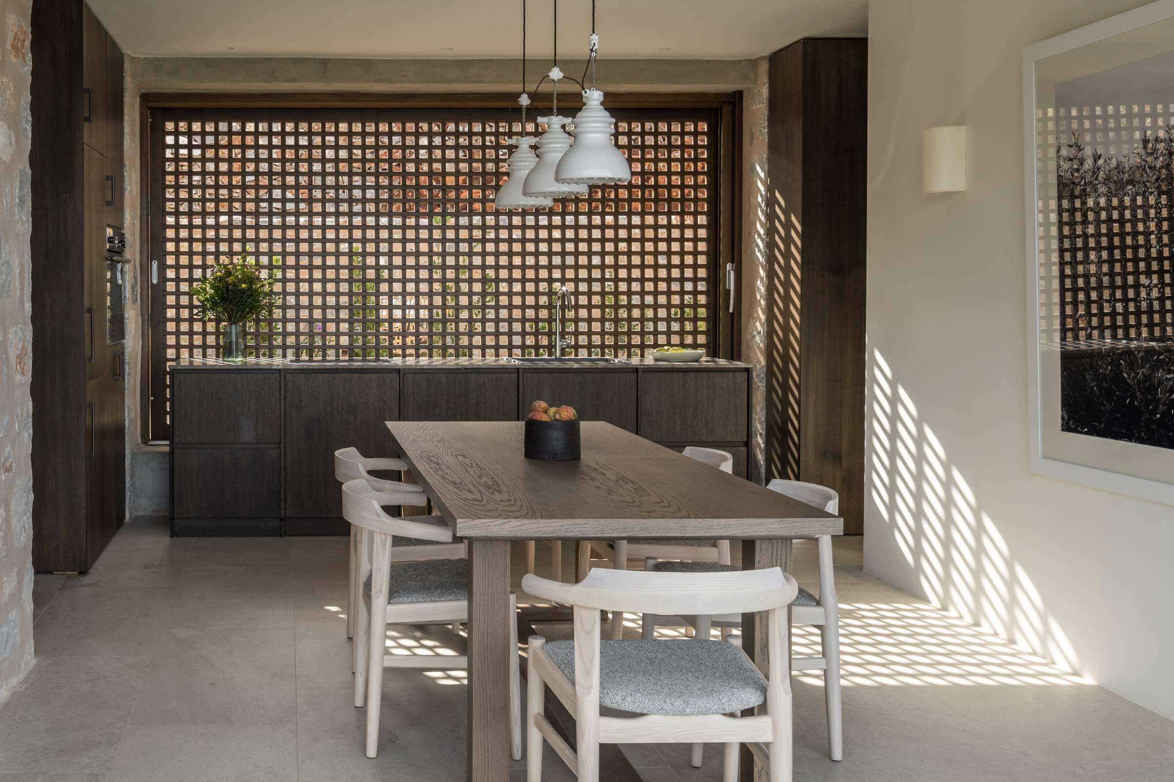 Monolithoi, Europe — Light-filled dining room with whitewashed wood table, lattice wall screens, pendant lights, and geometric floor patterns casting shadows.