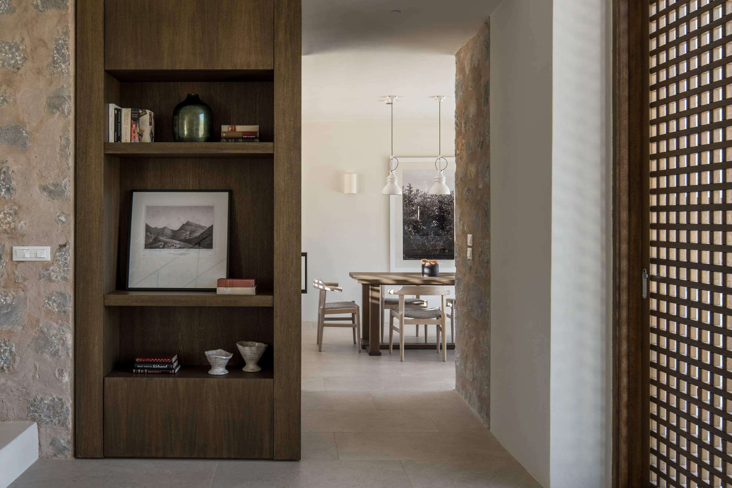 Monolithoi, Europe — Timber shelving unit with framed landscape photography, ceramic vessels, and books in minimalist hallway with stone walls and pendant lighting.