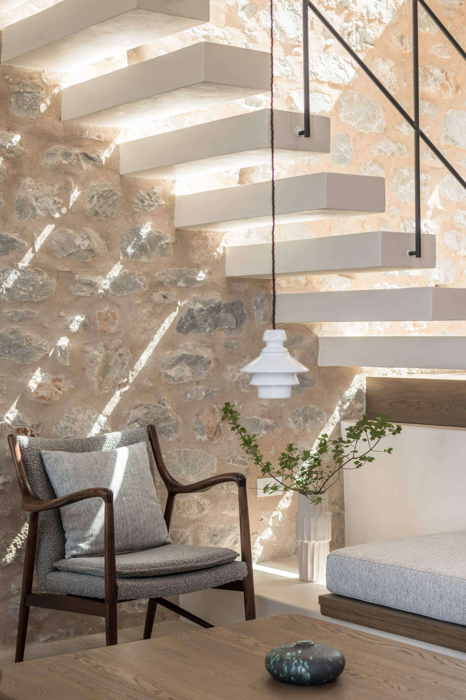 Monolithoi, Europe — Floating stone staircase with integrated lighting beside mid-century lounge chair and textured stone wall in minimalist interior.