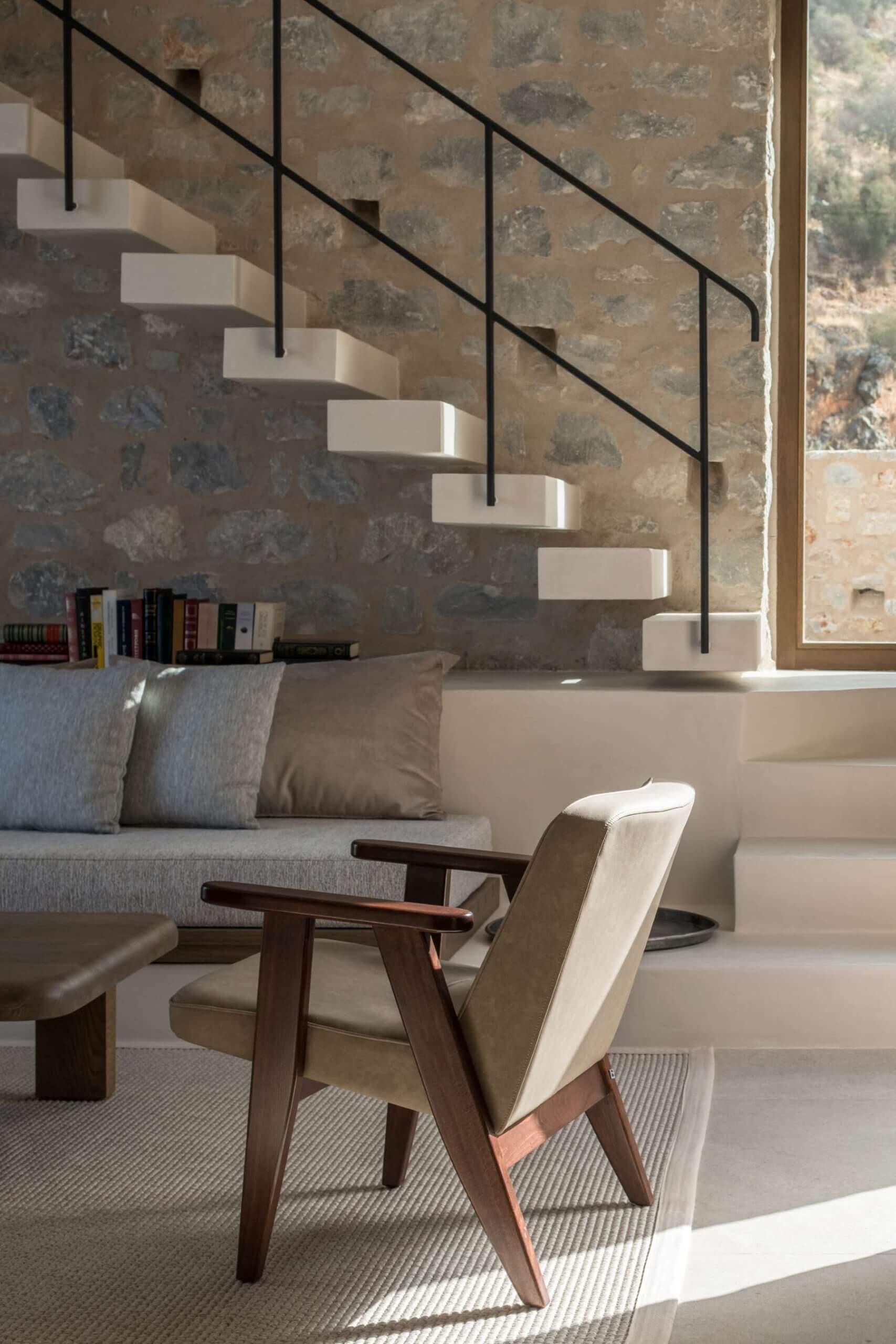 Monolithoi, Europe — Floating cantilevered staircase with black metal railings, weathered stone wall, mid-century modern chair, concrete steps, natural light, minimalist interior.