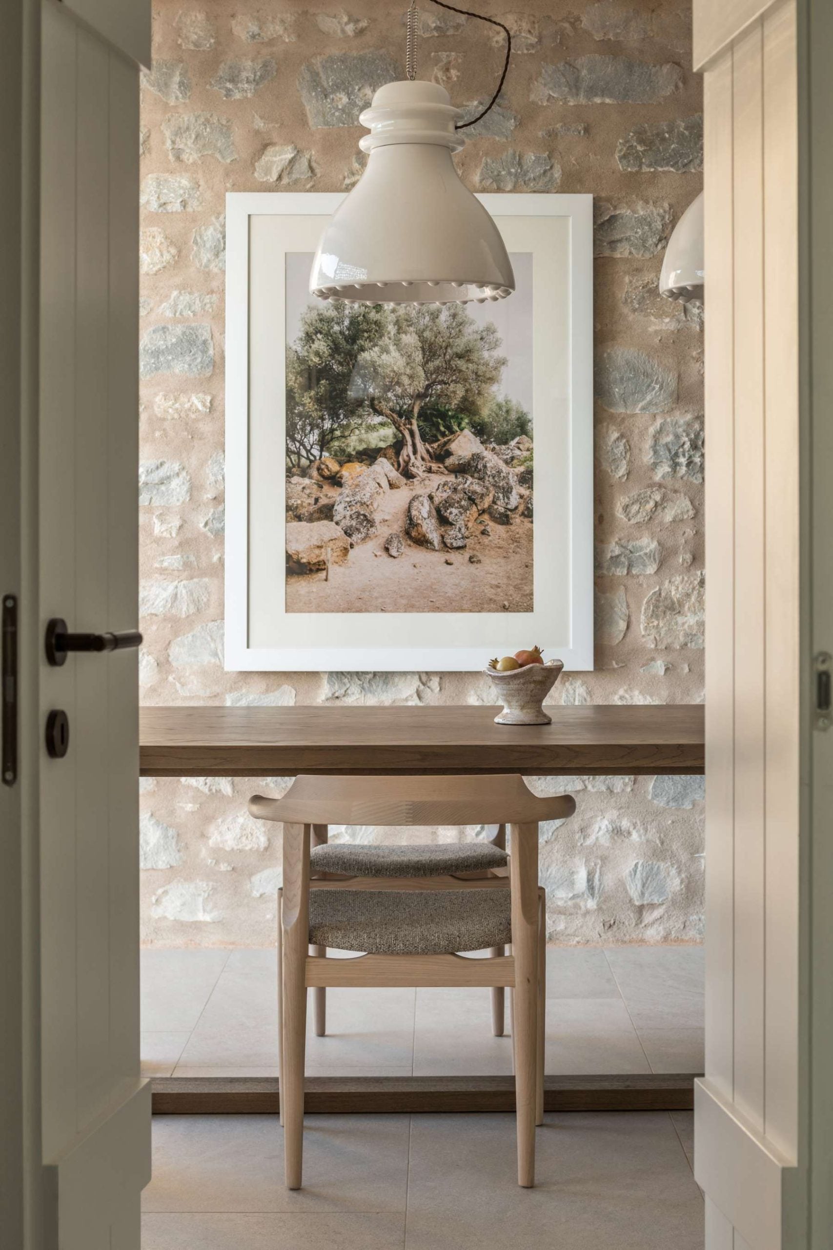 Monolithoi, Europe — Minimalist entryway with limestone brick wall, white pendant light, framed landscape photograph, natural wood desk and chair, neutral palette.
